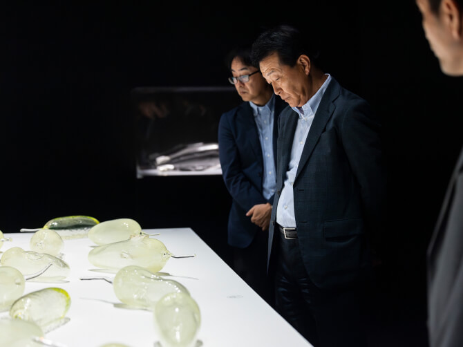 イワニ・スケースのウランガラス作品を鑑賞する参加者たち。暗い展示室で半透明のガラス作品が白い卓上に並ぶ。