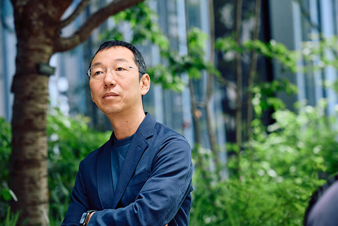 ミュージアムタワー京橋の緑の木々に囲まれた空間で、山口周氏が屋外で腕を組み、遠くを見つめている様子。