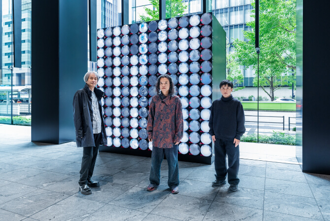 ミュージアムタワー京橋1階のエントランスに設置された《WINDOWS》の前で、左から高谷史郎氏、古舘健氏、濱哲史氏が真剣な表情で並んで立つ写真。ダムタイプのインタビュー記事「“変化するアートコレクティブ”の成り立ち」のサムネイル。